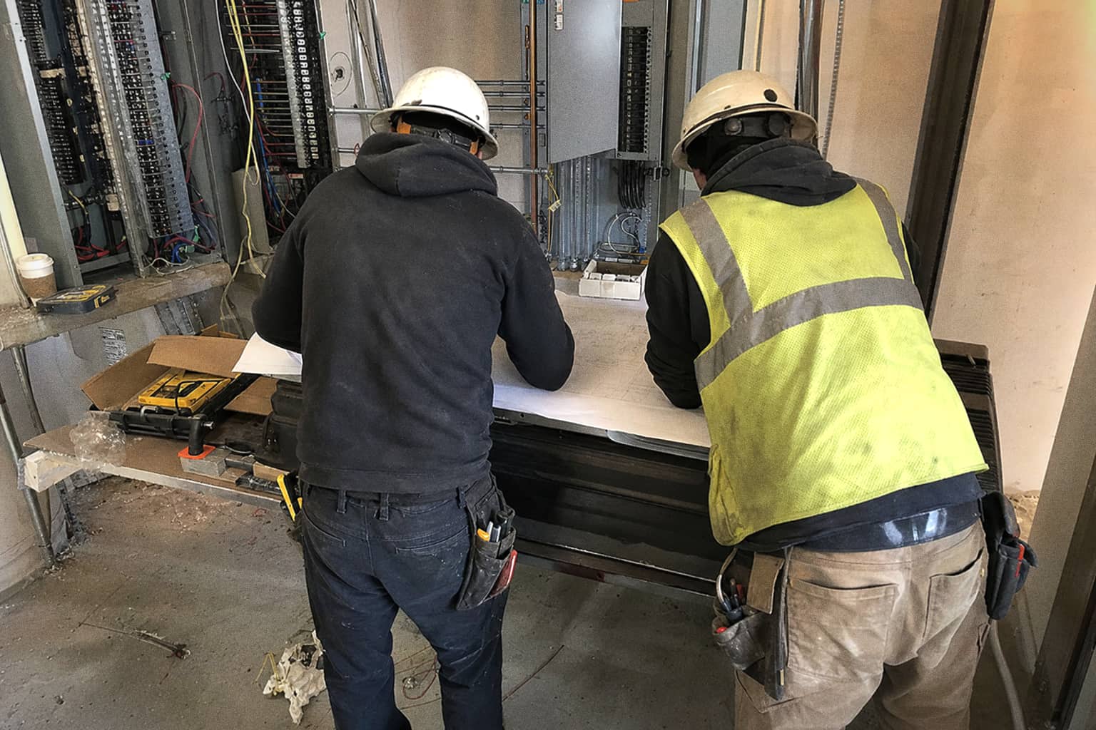 Two electricians reading a set of plans on a bench in front of open panelboards.